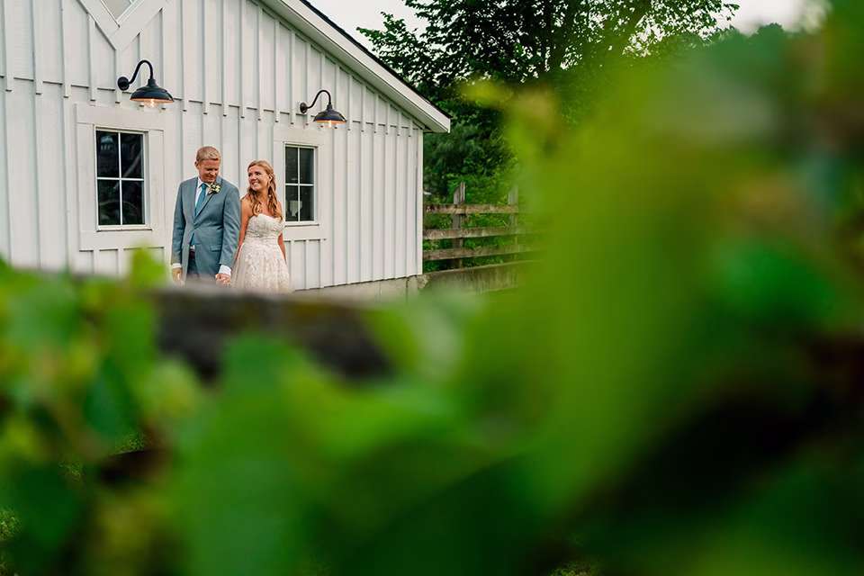 A bride and groom holding hands walk outside near the white Barn at Copper Creek, partially obscured by greenery in the foreground.
