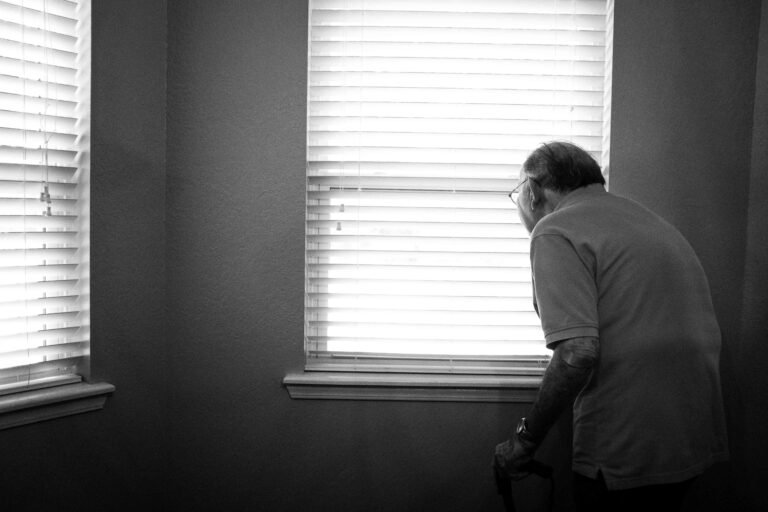 Older man standing at a window with blinds seen from behind, a caregiver’s view of a parent after a stroke