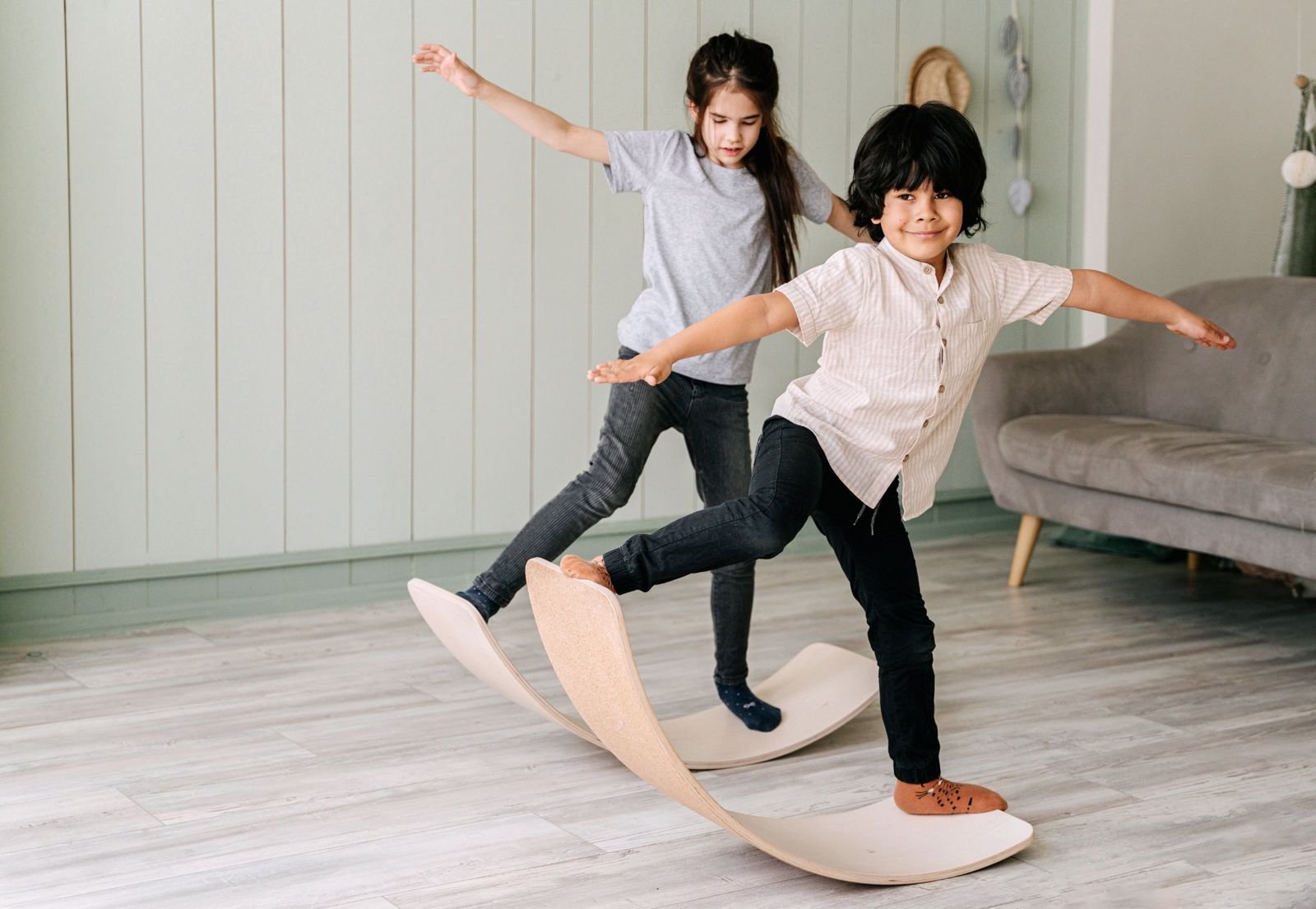 Two kids balancing with arms out on a curved wooden balance board indoors