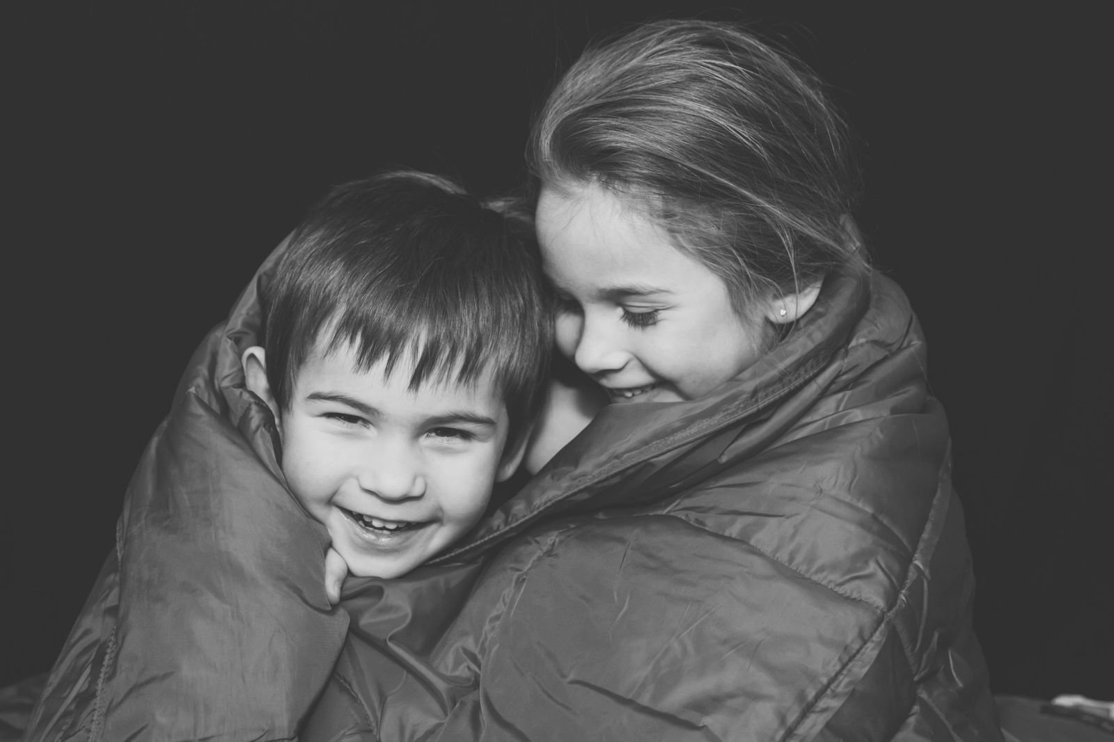 Two smiling children snuggled together inside a thick quilted blanket