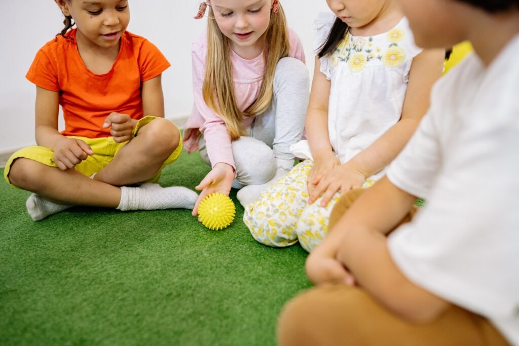 Children sitting on a green mat, one rolling a yellow spiky sensory ball across the floor