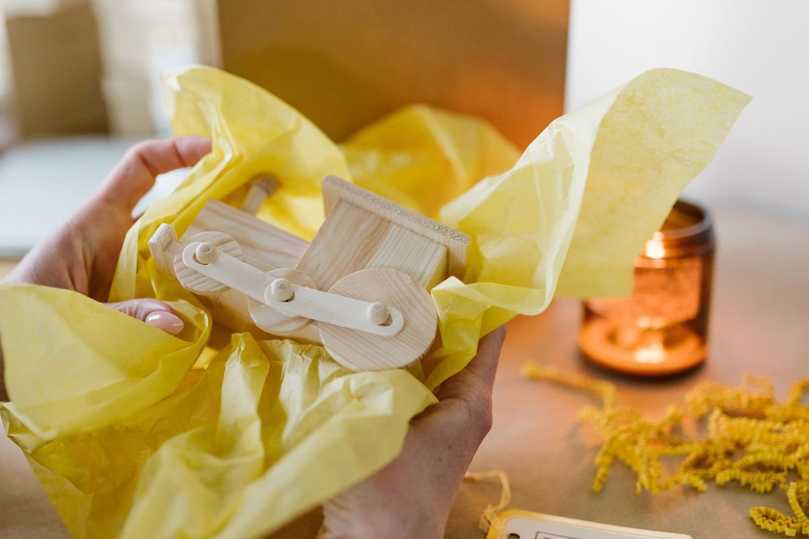 Hands holding a wooden toy in yellow tissue paper beside a plain kraft gift bag