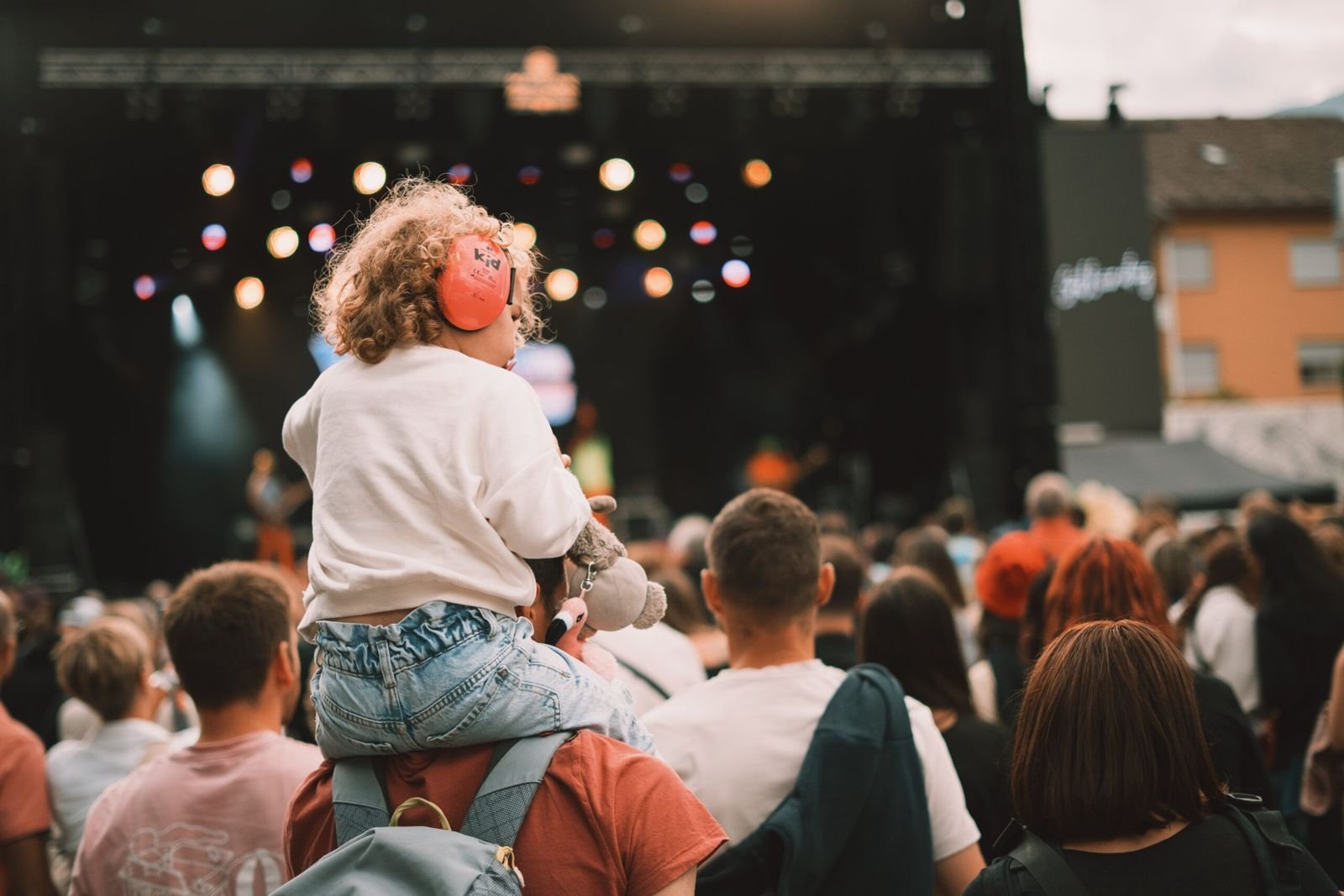 Toddler wearing red kids’ ear defenders on a parent’s shoulders at an outdoor concert
