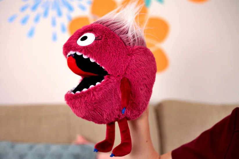 Colourful puppet with big eyes and open mouth, held up against a decorated background.