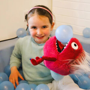 Child playing with a red plush fish puppet in a colourful ball pit.
