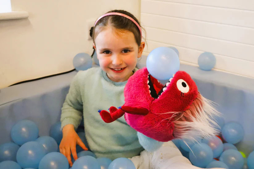 Child playing with a red plush fish puppet in a colourful ball pit.