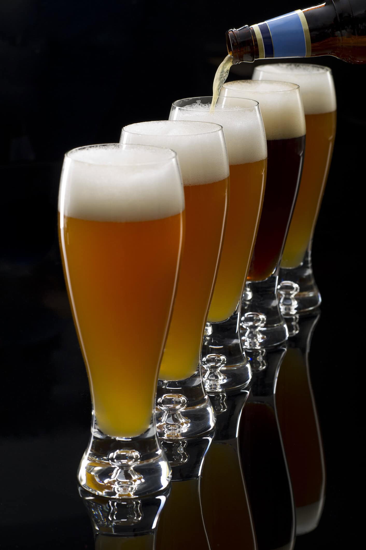 Five beer glasses with a bottle of beer being poured into the glasses.