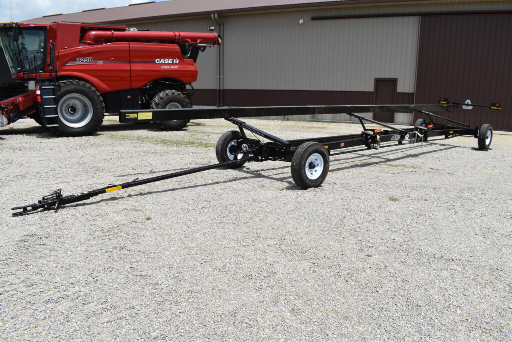 Farm equipment transport trailer on gravel with a red combine harvester in the background.