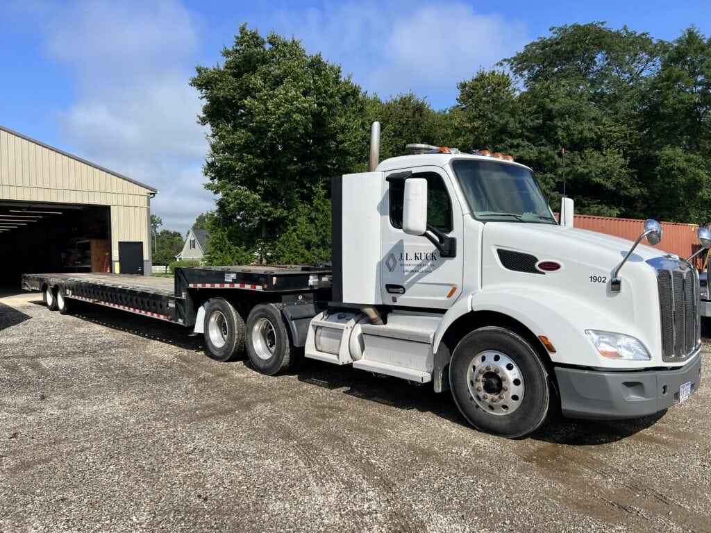 White flatbed truck at auction site with trees and building in background.
