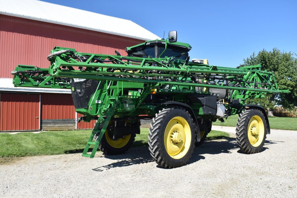 Green John Deere agricultural sprayer with large tires and spray arms.
