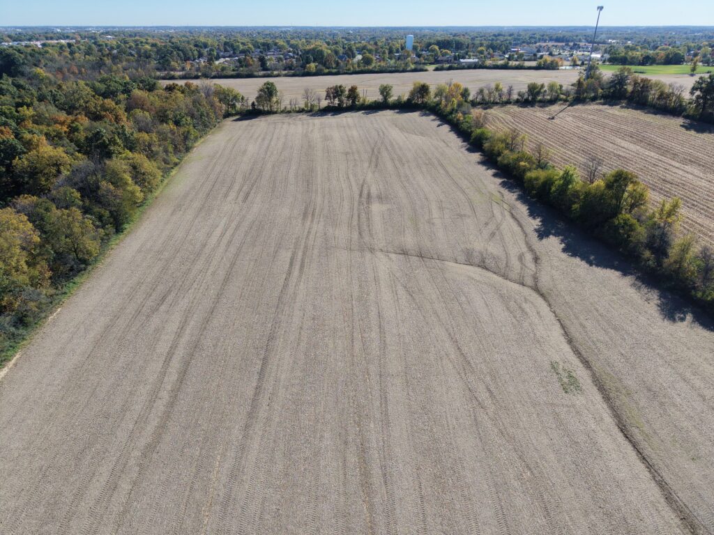 Aerial view of a large, open farmland field with surrounding trees and distant cityscape.