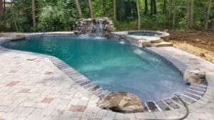 A pool with a waterfall feature surrounded by a stone patio in a wooded backyard.