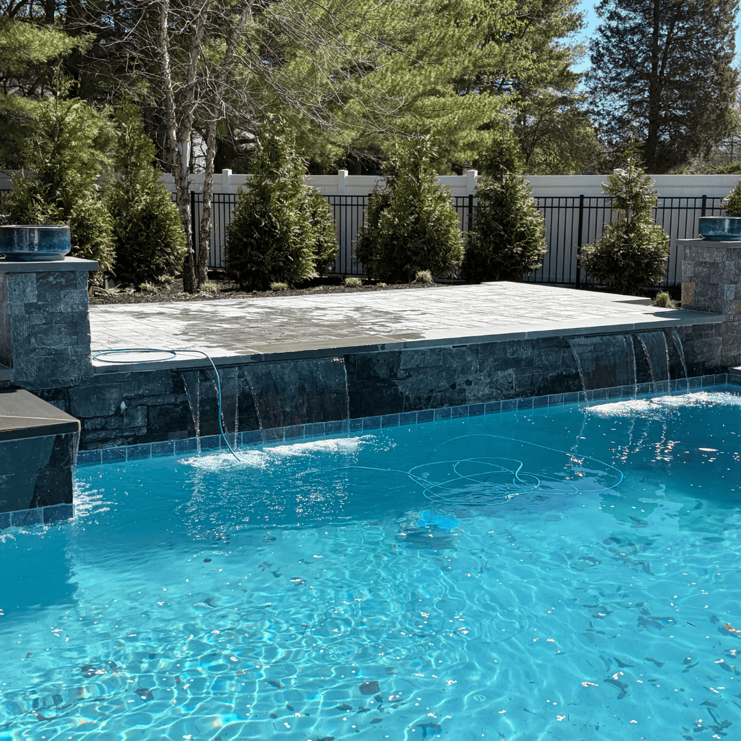 Backyard swimming pool with waterfall and stone deck, surrounded by lush trees and privacy fence.