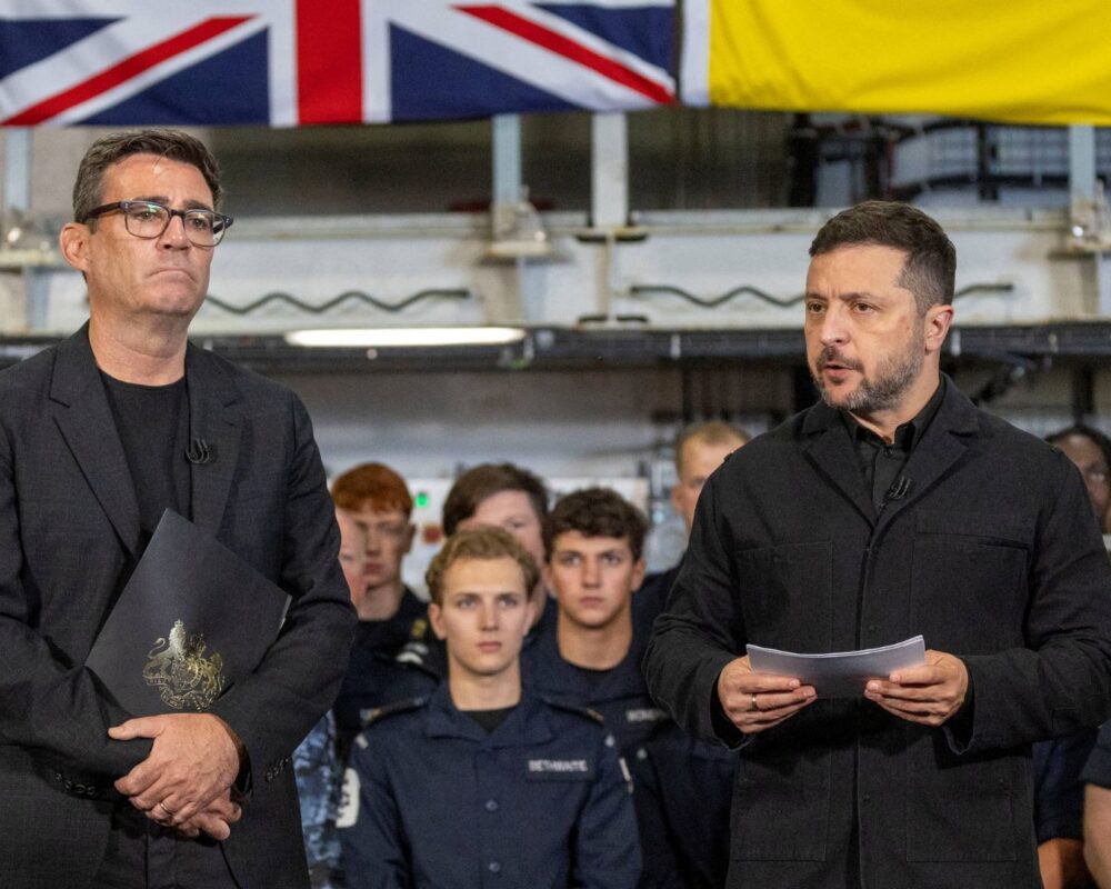 Military officials and personnel at a briefing with UK and yellow flags in background.