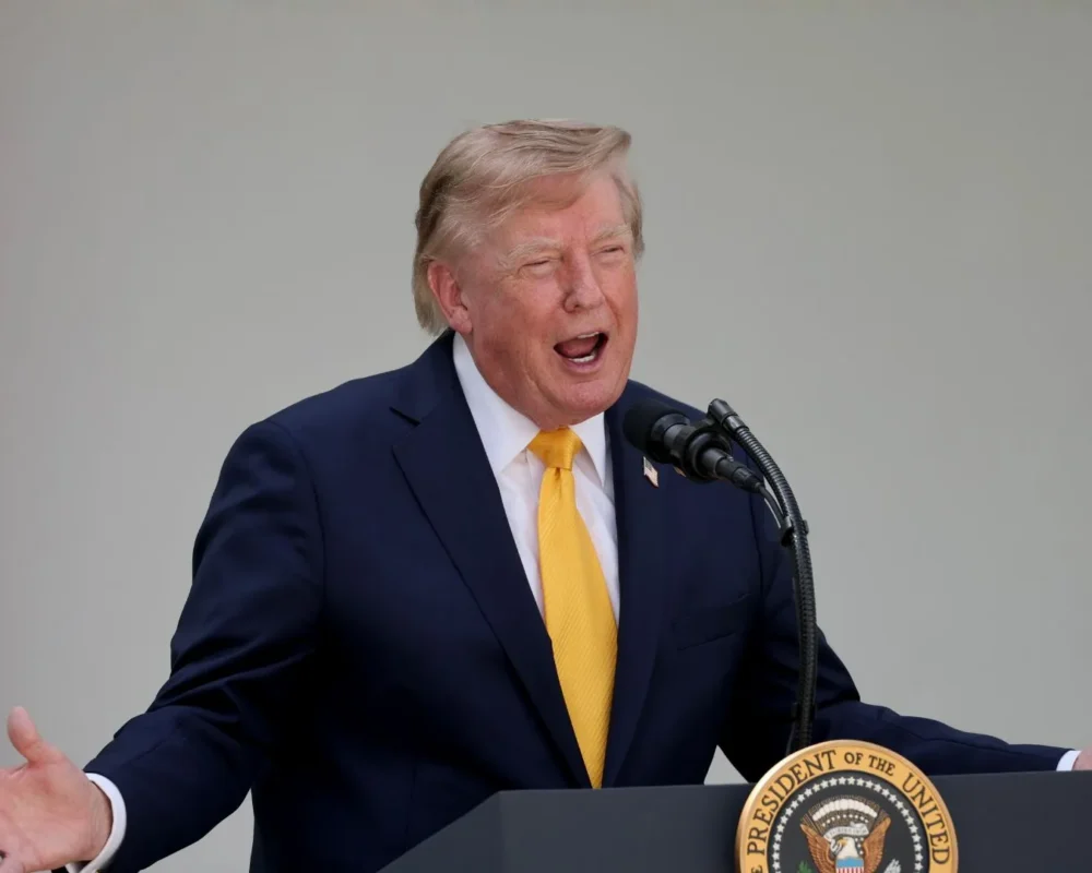 Donald Trump speaking passionately at a press event with American flag in background.