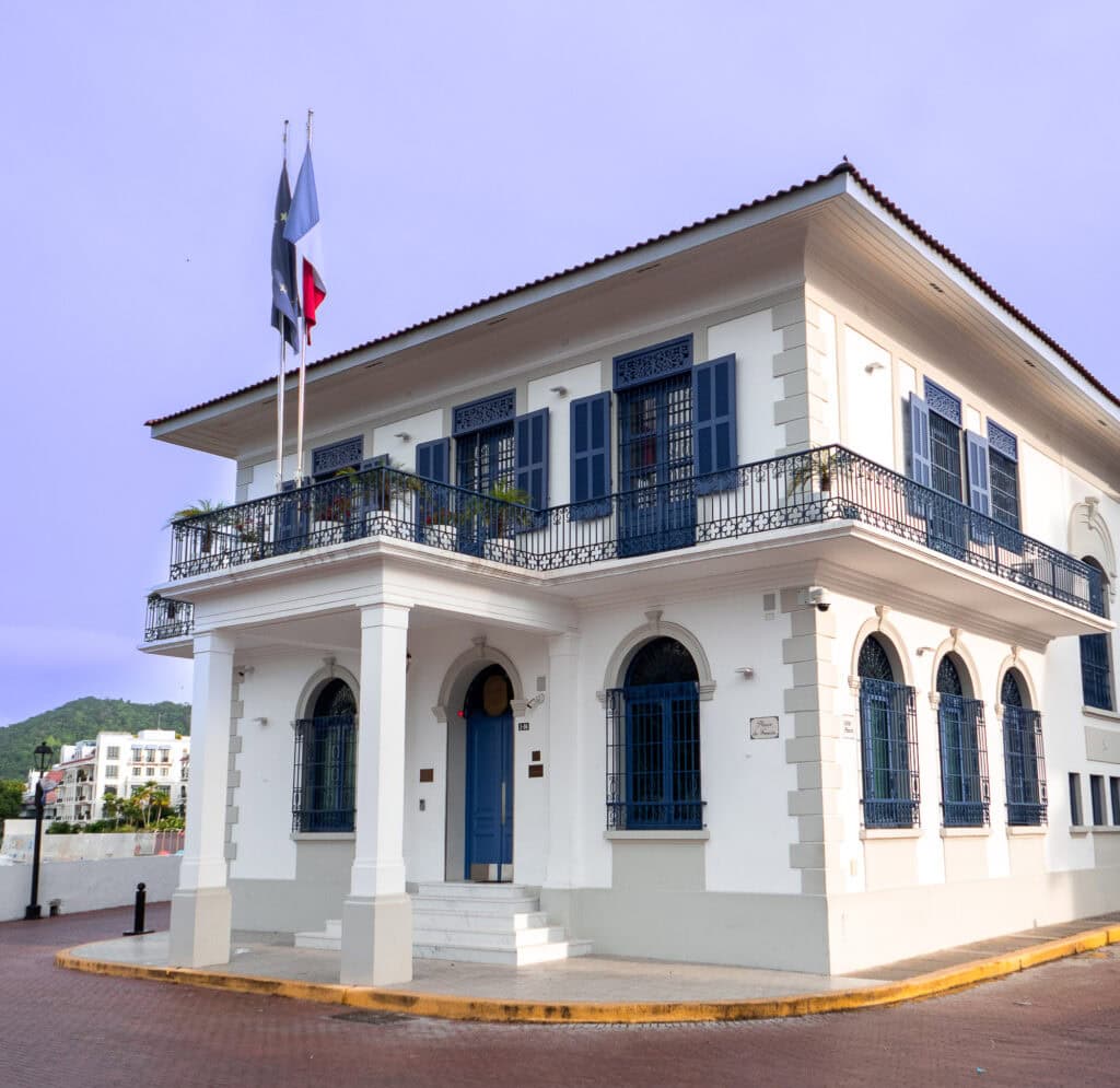 Elegant white building of the French Embassy in Casco Viejo, Panama, with blue shutters and French and Panamanian flags flying above the main entrance.
