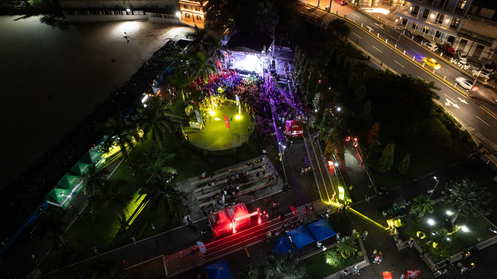 Aerial night view of Casco Viejo Pride concert with colorful lights, a beachfront crowd, palm trees, and a lit-up main stage in Panama City.