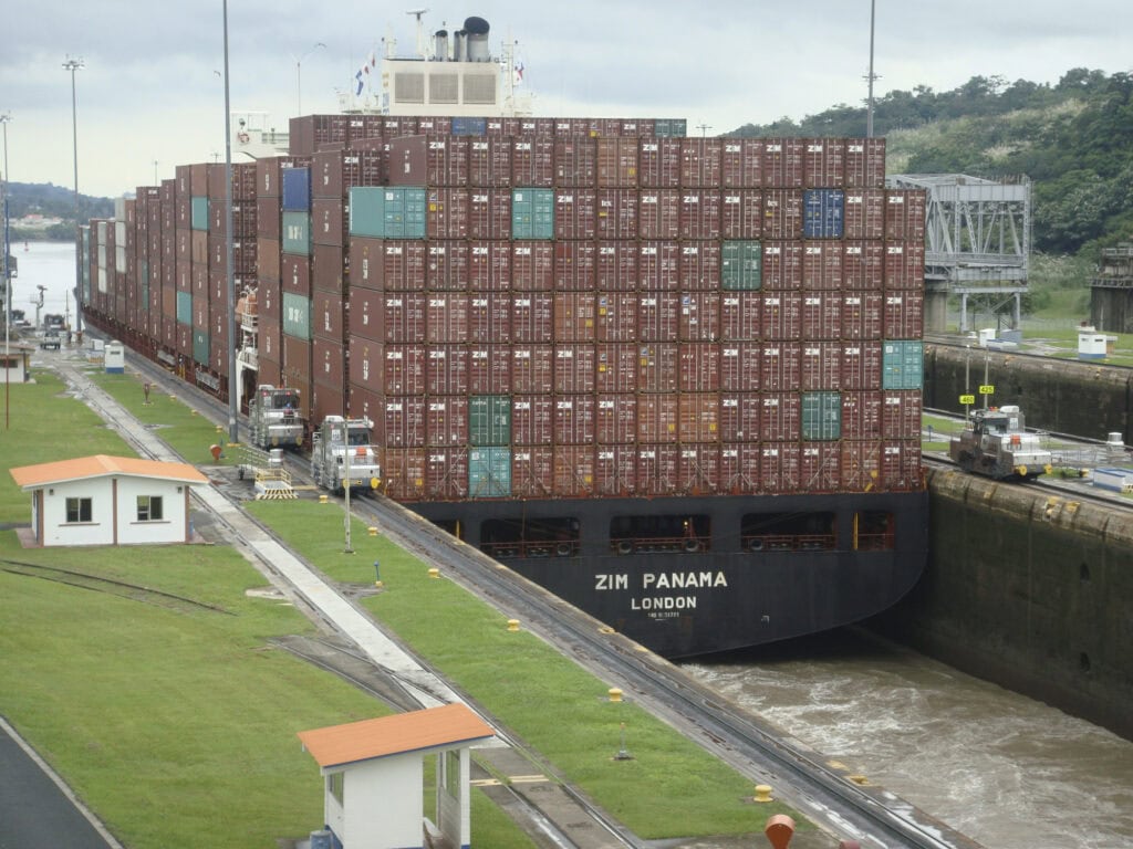 A container ship named ZIM Panama navigates the narrow Mira Flores Locks of the Panama Canal, guided by locomotives on either side.