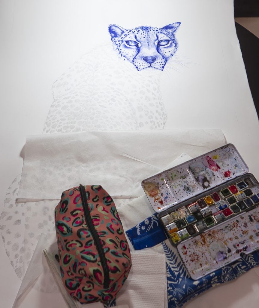 Partially finished watercolor painting of a cheetah with an artist's paint palette and patterned pencil case beside it
