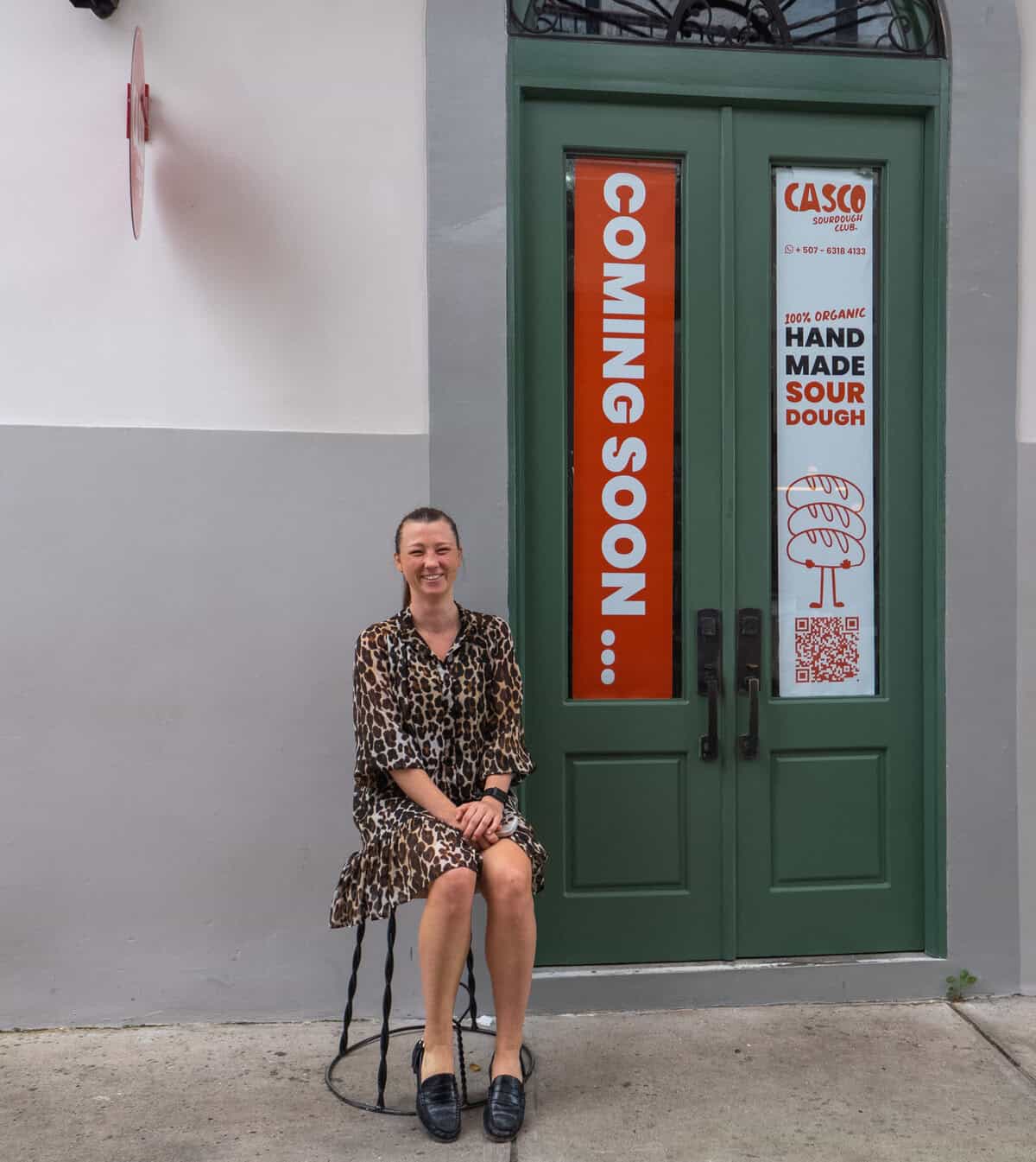 Caroline sitting on a stool outside her workspace, smiling in front of a green door.