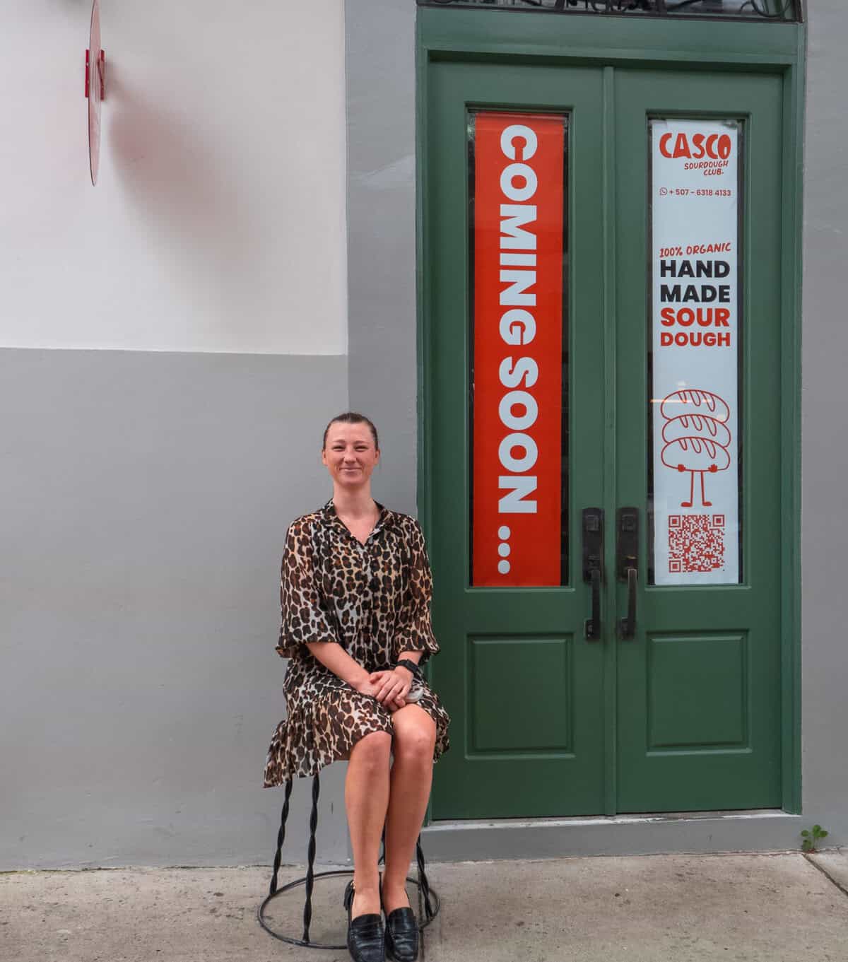 Caroline seated casually outside, smiling in front of tall green doors with an orange banner beside her.