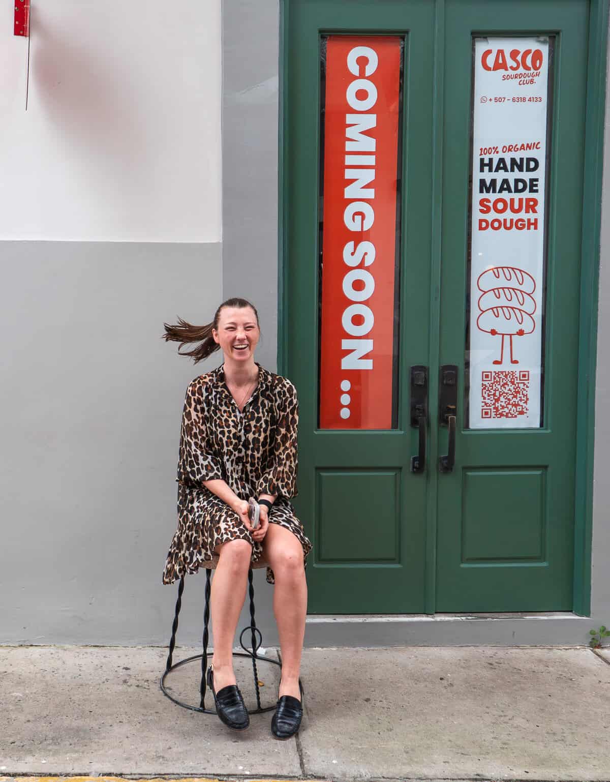 Caroline laughing as a breeze lifts her hair while she sits on a stool outside near the green doors.
