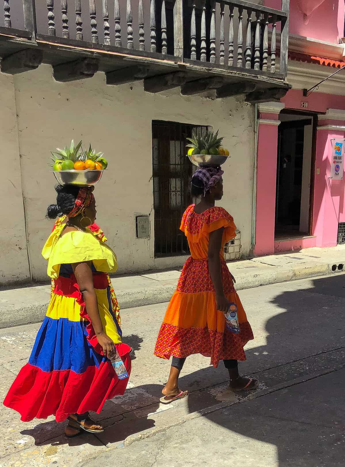 Two Palenquera women in colorful traditional dresses carrying fruit through Cartagena’s historic walled city.