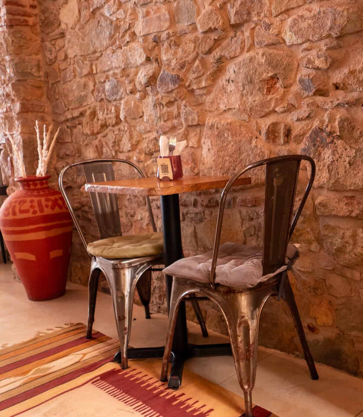 Table for two with cushioned metal chairs beside an exposed stone wall and a large red decorative vase at The Vegan Shack.