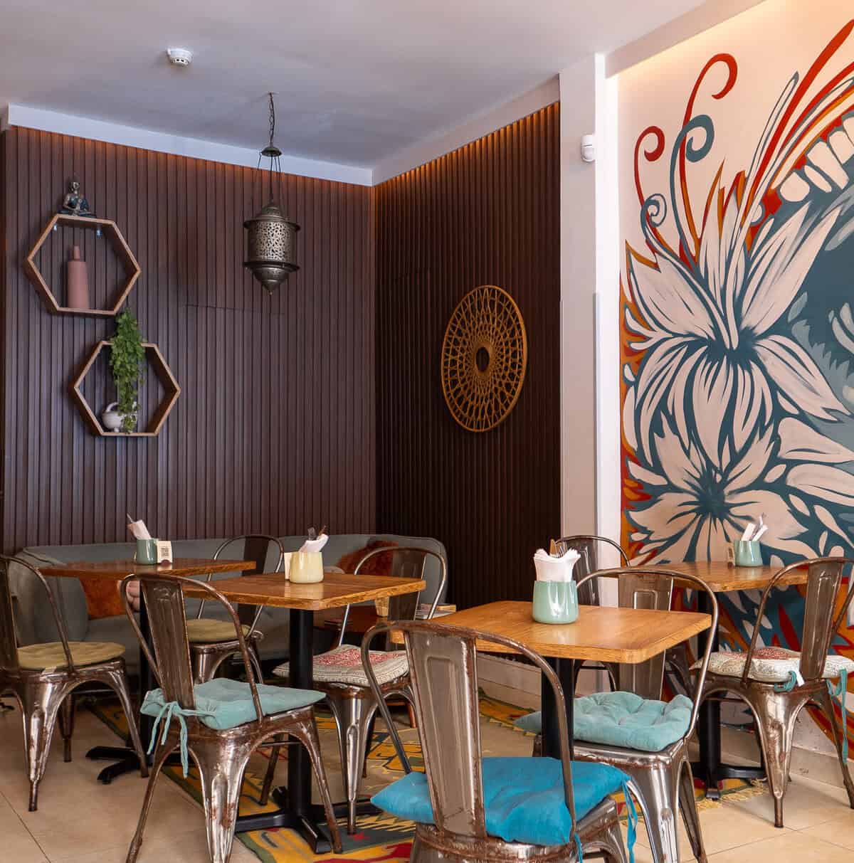 Wooden tables and metal chairs with colourful cushions beside a floral mural inside The Vegan Shack in Casco Viejo.