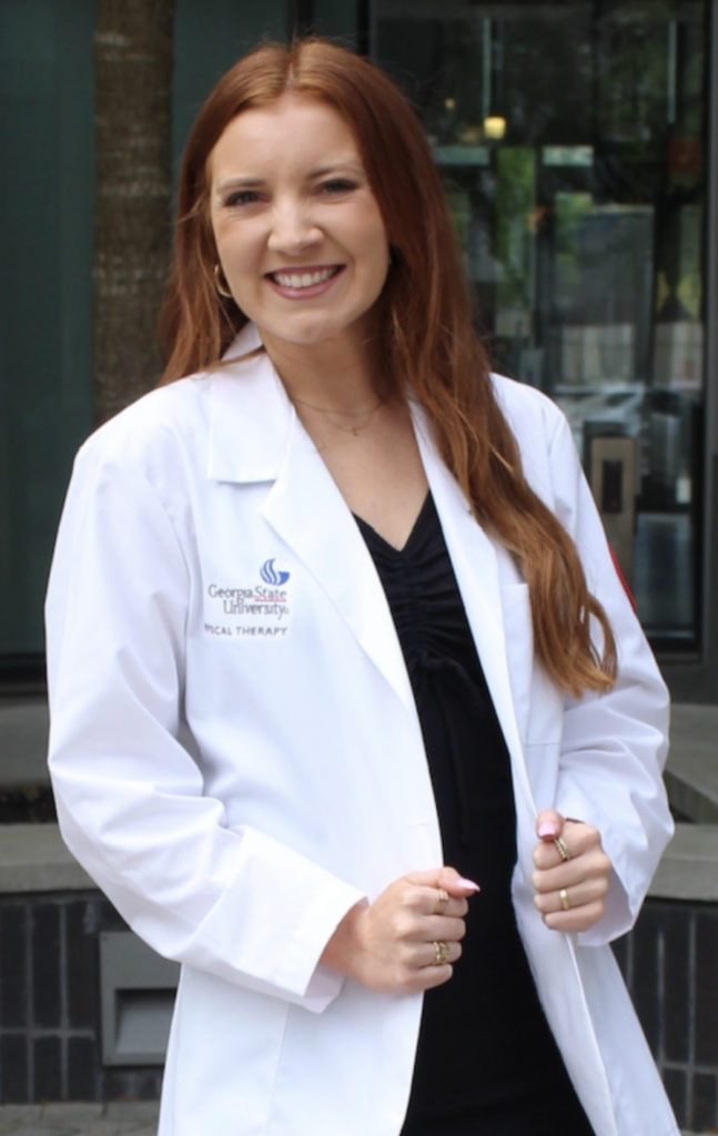 A woman with long red hair wearing a white Georgia State University Physical Therapy lab coat stands outdoors in Atlanta, smiling at the camera—ready to make a difference in pediatric physical therapy.