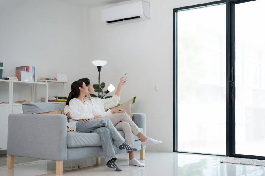 Couple relaxing on a sofa while adjusting a Daikin wall-mounted air conditioner with a remote.
