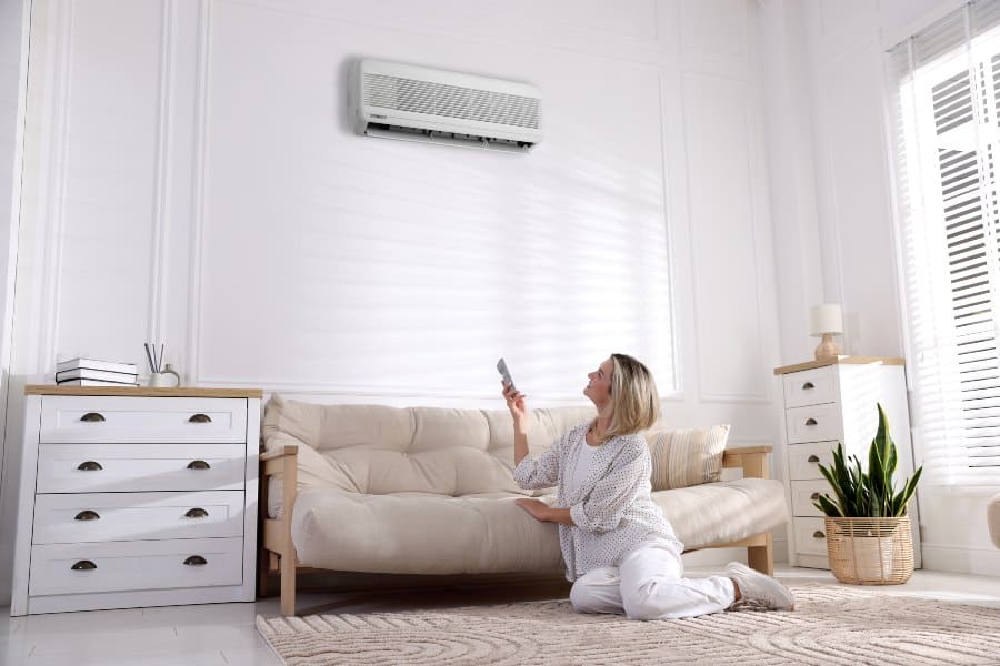 Woman adjusting a split system air conditioner with a remote in a bright living room.
