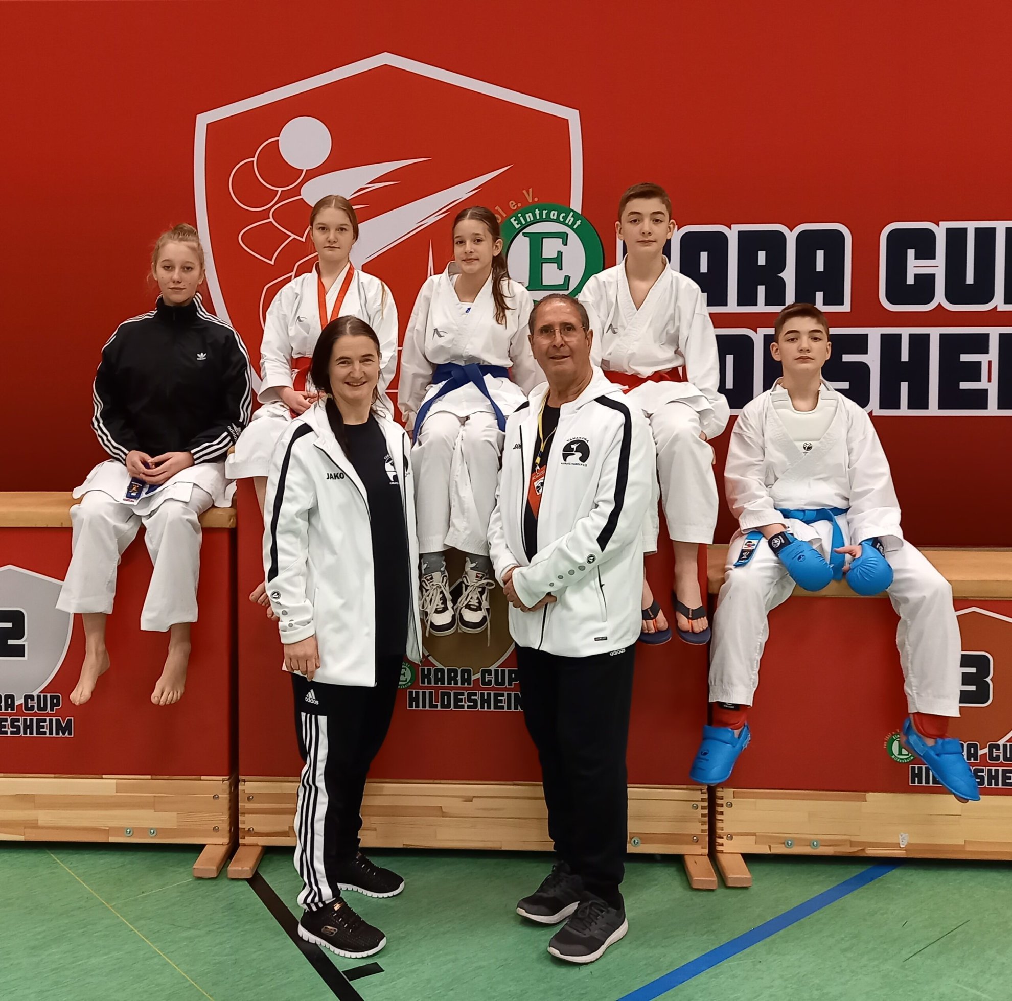 Karate-Team posiert in Sportbekleidung vor rotem Banner.