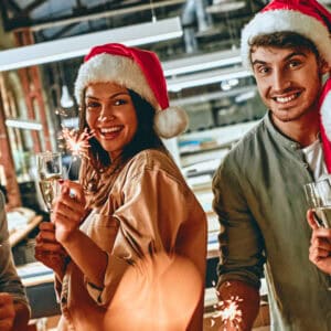 Vier lachende junge Menschen mit Weihnachtsmützen feiern mit Sektgläsern und Wunderkerzen
