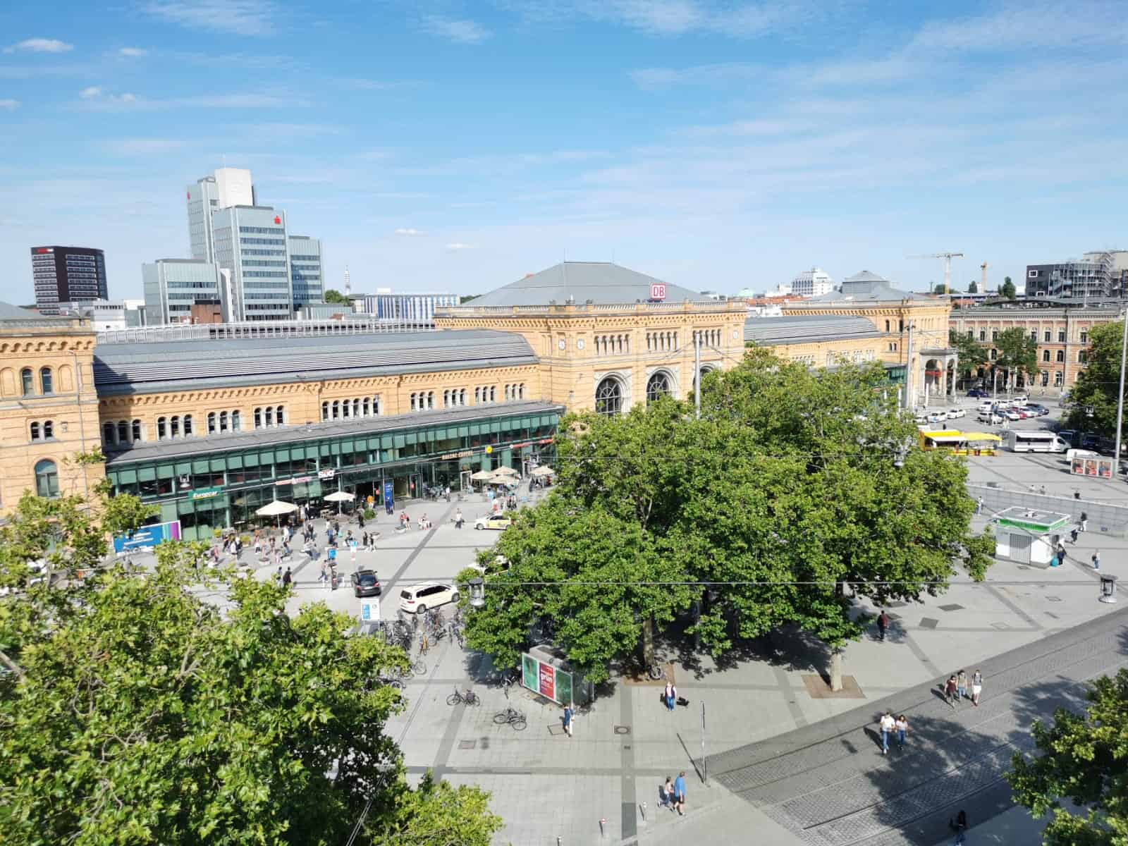 Blick von einem oberen Geschoß des Hotels auf den Ersnt-August-Platz mit grünen Bäumen und den Bahnhof. 