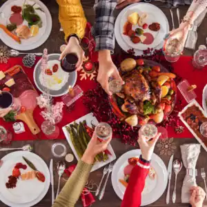 Weihnachtlich gedeckter Tisch von oben mit gebratenem Geflügel in der Mitte, umgeben von Tellern mit Aufschnitt, Lachs, Spargel und Salat; mehrere Personen stoßen mit Wein- und Sektgläsern über der Festtafel an