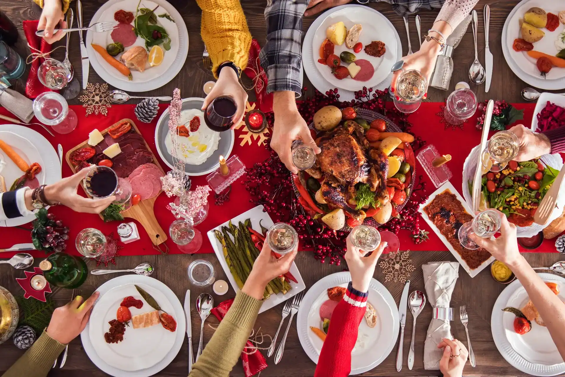 Weihnachtlich gedeckter Tisch von oben mit gebratenem Geflügel in der Mitte, umgeben von Tellern mit Aufschnitt, Lachs, Spargel und Salat; mehrere Personen stoßen mit Wein- und Sektgläsern über der Festtafel an