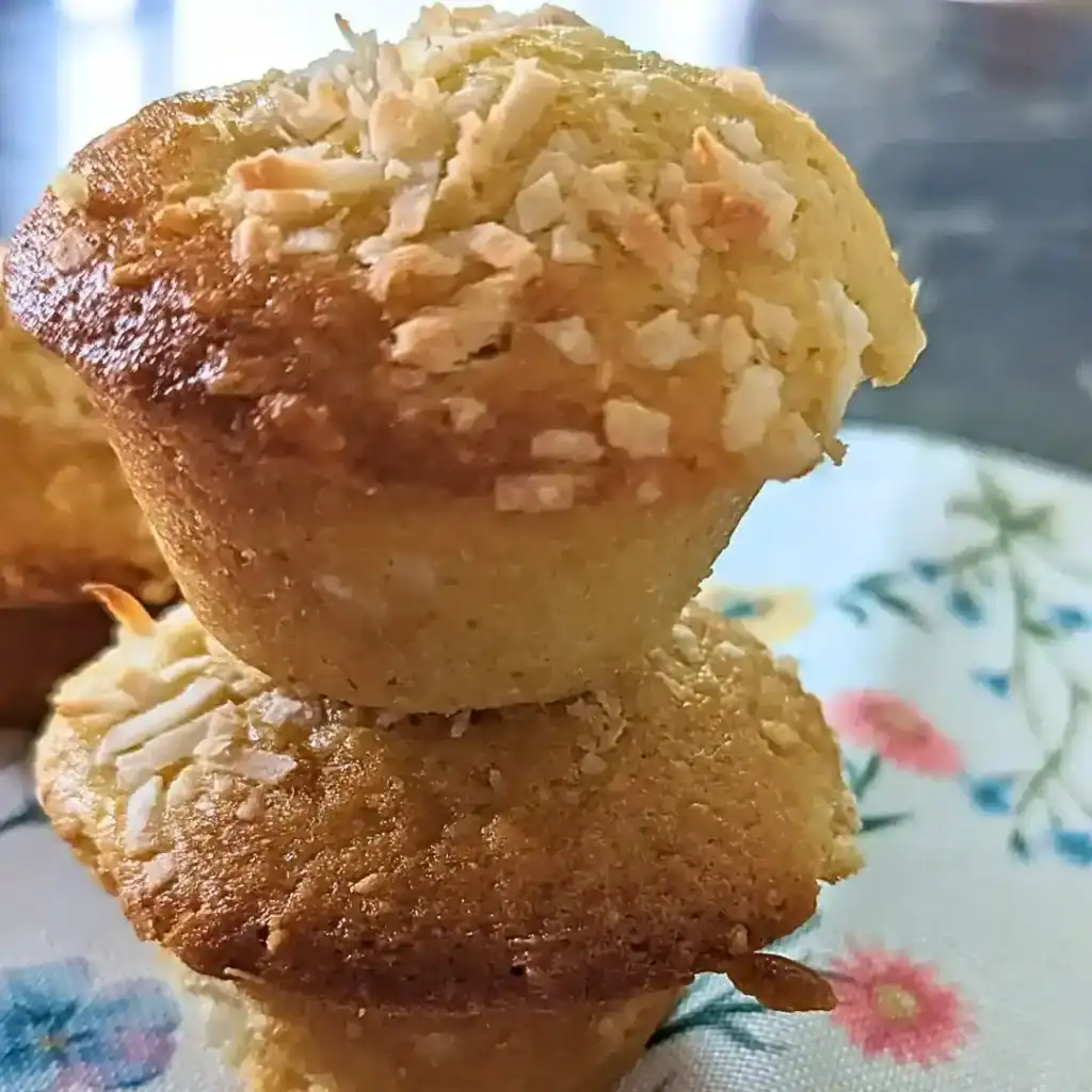 the recipe card image of a peach coconut muffin stacked on another muffin on a floral placemat background blurred out