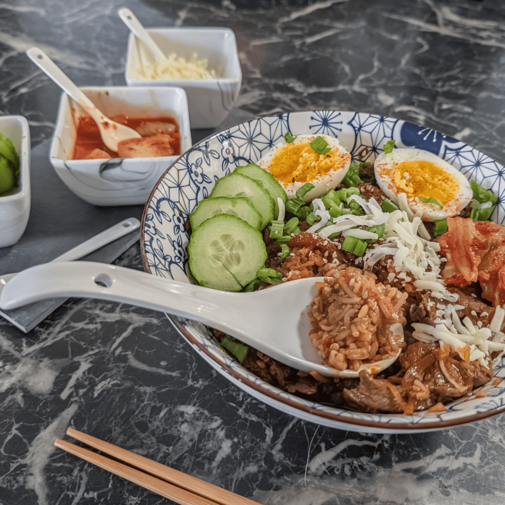 Close-up of a Japanese beef rice bowl with eggs, cucumbers, and toppings on a dark marble table.