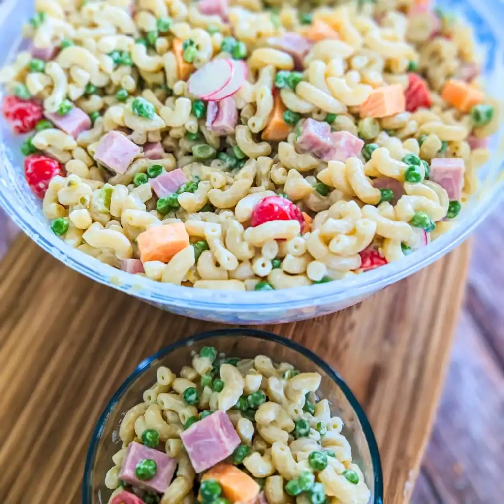 creamy macaroni salad with ham and peas side of large plastic bowl on wood cutting board top down view and smaller serving bowl