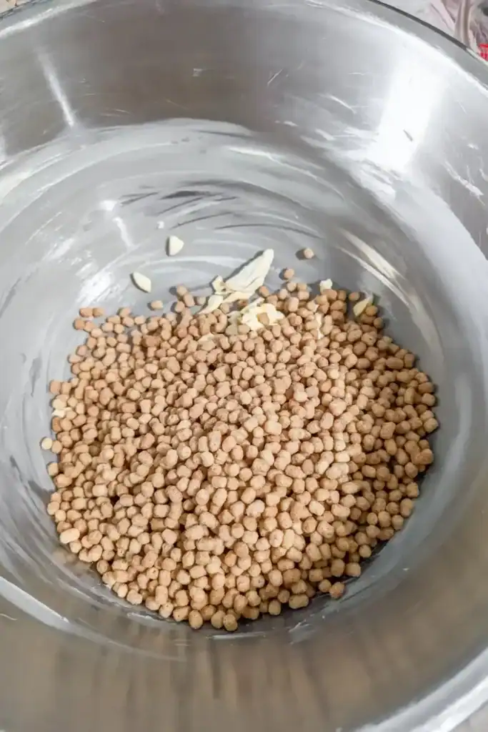 adding mini cinnamon toast crunch cereal to dried apple in buttered stainless steel mixing bowl