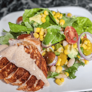 Fresh grilled chicken salad with corn, cherry tomatoes, and greens on a white plate.