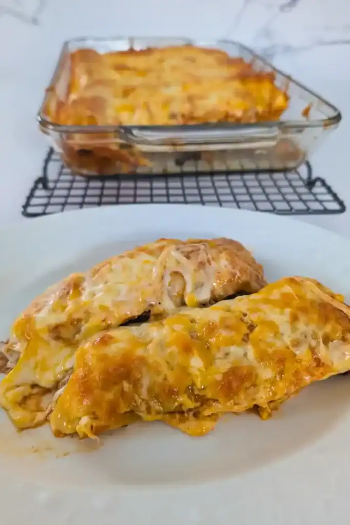 two cheesy ground chicken enchiladas on white plate foreground pan of enchiladas behind
