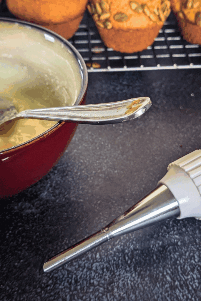 Buttercream frosting in a red mixing bowl with a piping tip and piping bag on a black countertop, baked muffins in the background.