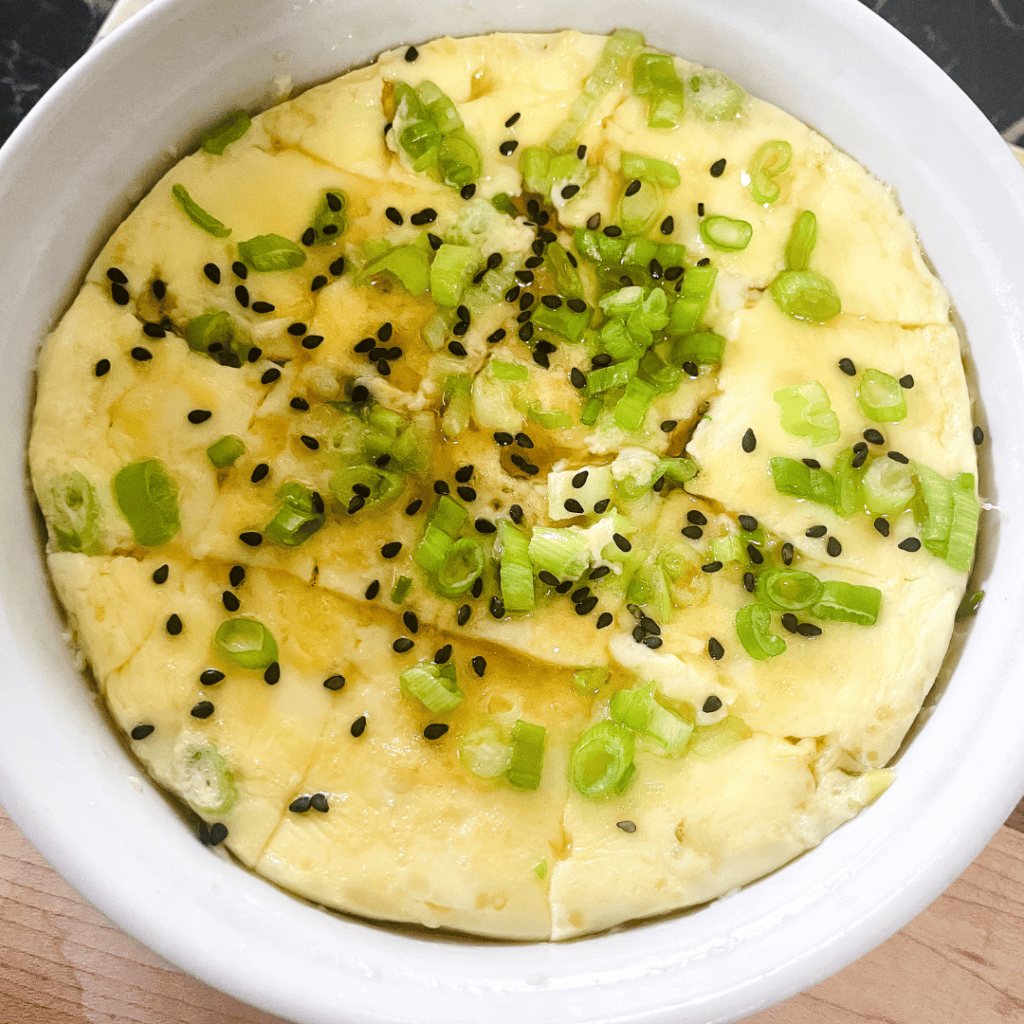Egg dish topped with chopped green onions and black sesame seeds in a white bowl.