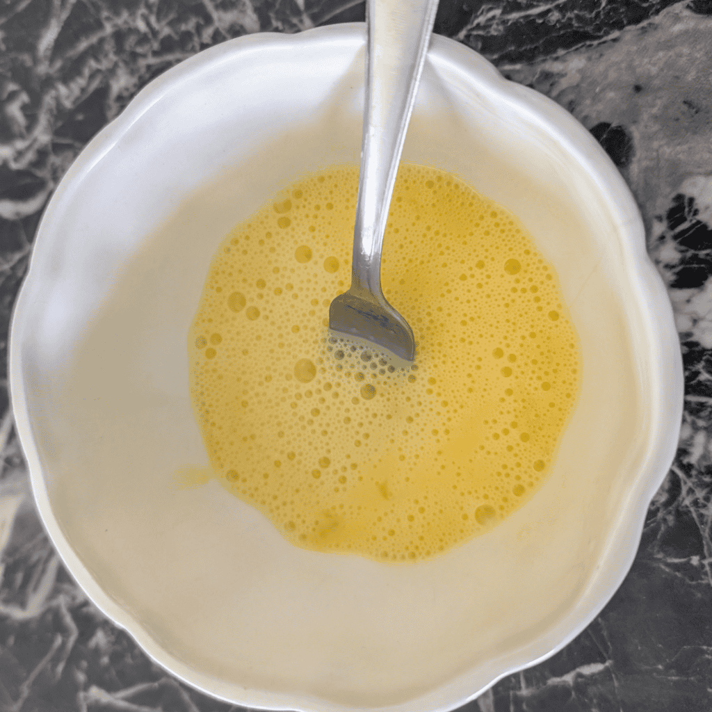 Freshly prepared egg wash in a white bowl with a spoon, ready for cooking or baking.