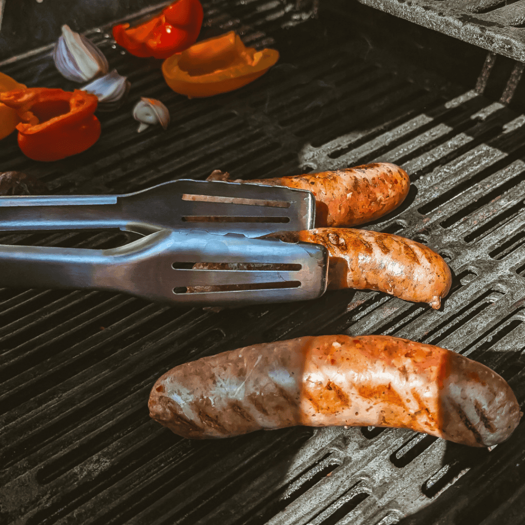 Close-up of sausages grilling on barbecue with peppers and garlic in the background.