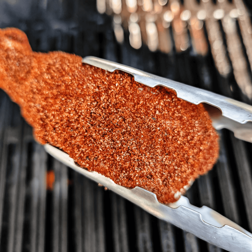 Close-up of seasoned BBQ chicken drumstick on grill tongs.