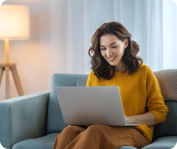 Woman in yellow sweater with laptop on sofa near lamp and curtains for remote work infrastructure.