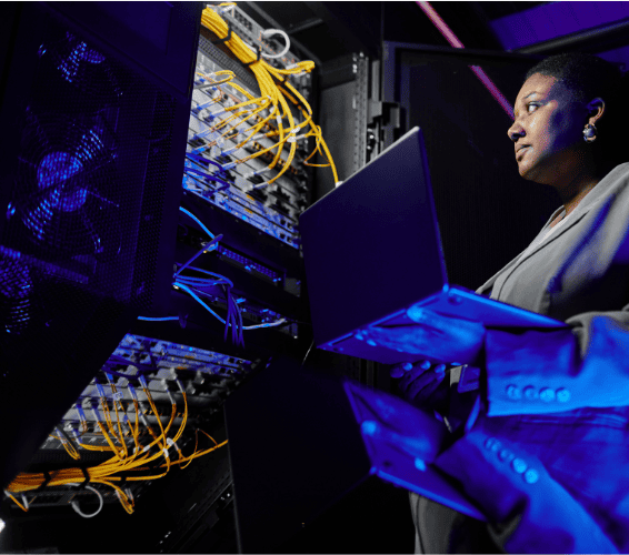 IT professional with a laptop beside yellow-cabled network servers in a dim server infrastructure environment.